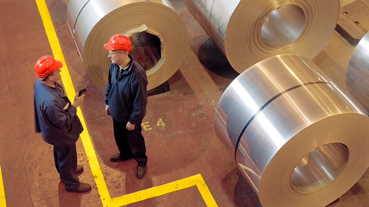 Two factory workers standing next to rolls of steel