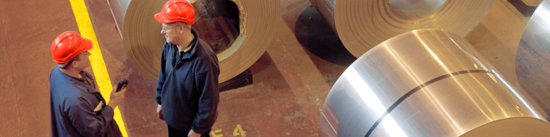 Two factory workers standing next to rolls of steel
