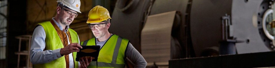 2 men in high vis jackets, hard hats and safety glasses in foreground, large industrial machinery in background