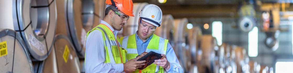 2 hommes en gilet jaune et casque de sécurité consultant une tablette, matériel industriel empilé à l’arrière-plan