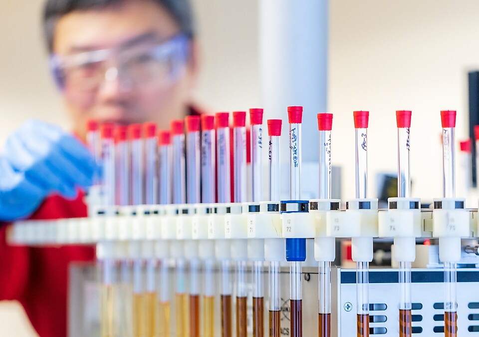 Technicien dans un labo, en blouse rouge et gants bleus, manipulant des tubes à essai contenant des échantillons de liquide