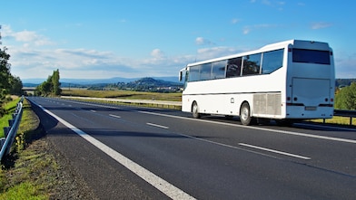 White Bus driving on an empty road in a rural landscape. Villages and forested mountains in the background.