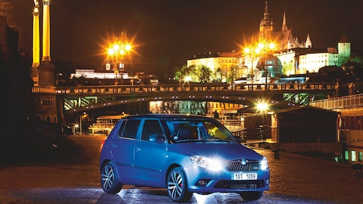 Une voiture bleue dans une rue de Prague avec un pont et un château en arrière-plan, de nuit