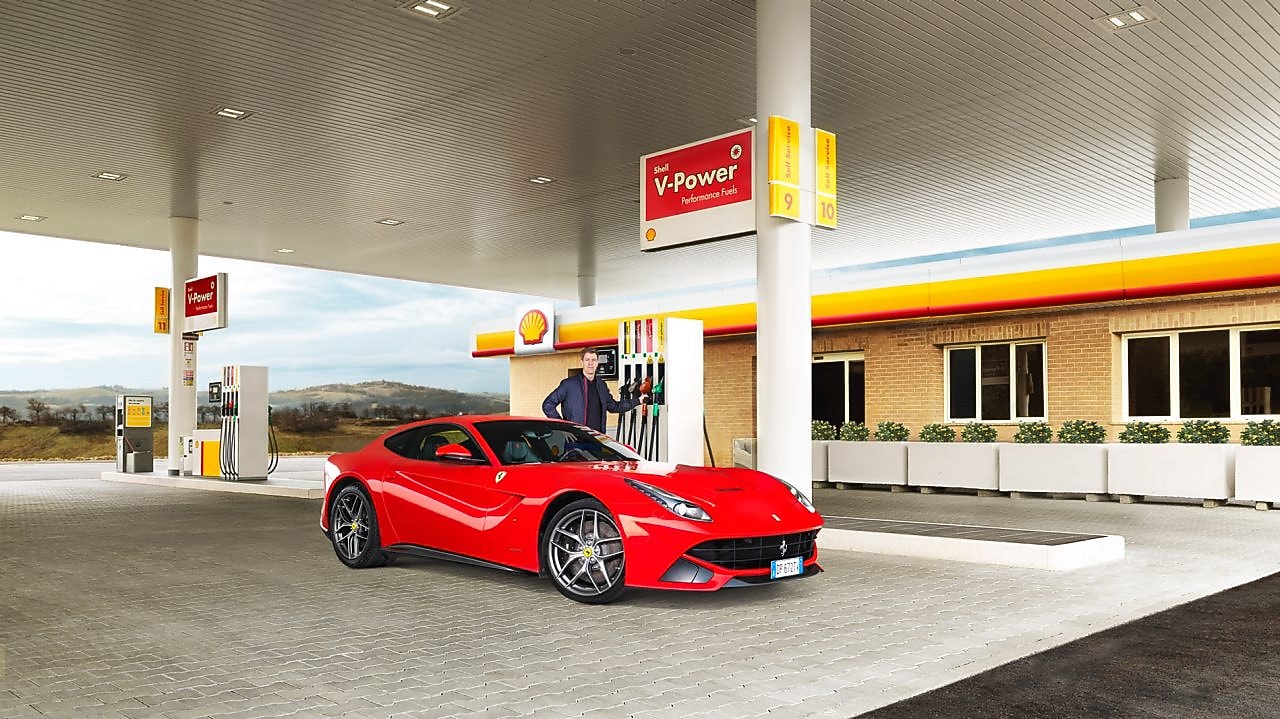 Une Ferrari rouge arrêtée à la pompe d'une station Shell avec un homme qui s'apprête à faire le plein