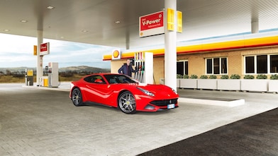 Une Ferrari rouge arrêtée à la pompe d'une station Shell avec un homme qui s'apprête à faire le plein