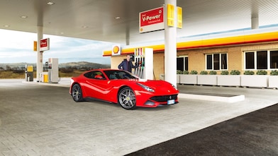 Une Ferrari rouge arrêtée à la pompe d'une station Shell avec un homme qui s'apprête à faire le plein